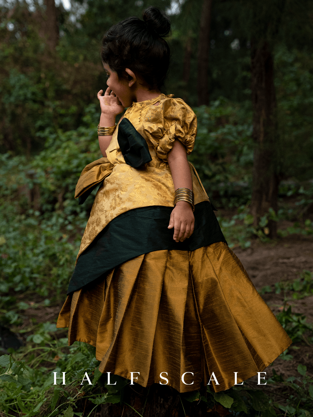 Grandeur Emerald Augustan Kanjivaram Silk Baby Girl Princess Dress with floral jacquard pattern, puff sleeves, and a twisted gold-green bow on the waistline.