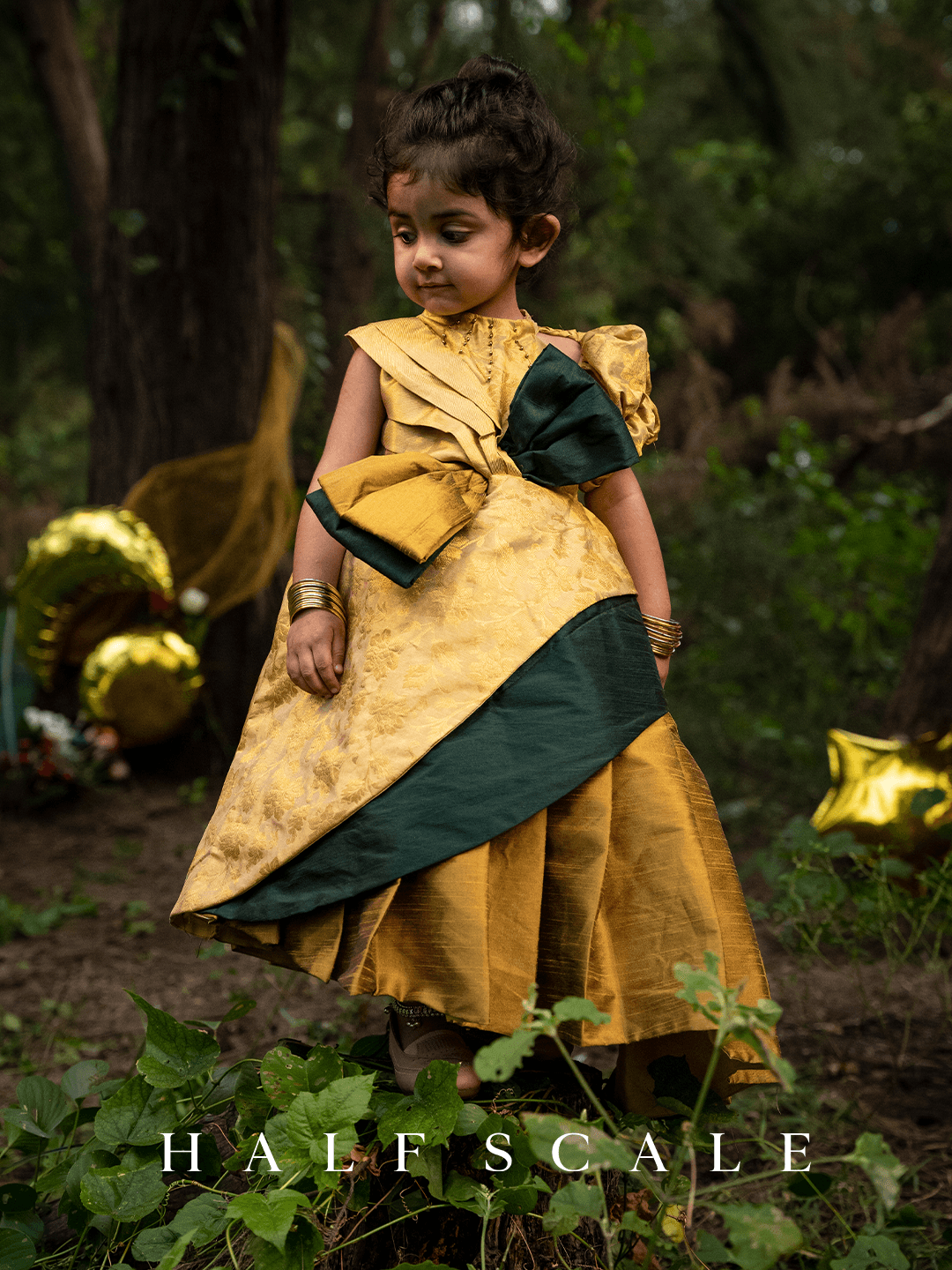 Grandeur Emerald Augustan Kanjivaram Silk Baby Girl Princess Dress with floral jacquard pattern, puff sleeves, and a twisted gold-green bow on the waistline.